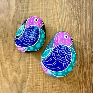 May include: Two hand-painted bird-shaped stones. Each stone features a pink head, a blue body with a detailed feather design, and teal accents. The birds are set against a wooden background, showcasing the vibrant colors and artistic detail.