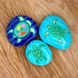 May include: Three painted rocks with turtle designs. The rocks are blue and turquoise, with white dotted borders. The turtles are green and have colorful shell patterns. The rocks are on a wooden surface.