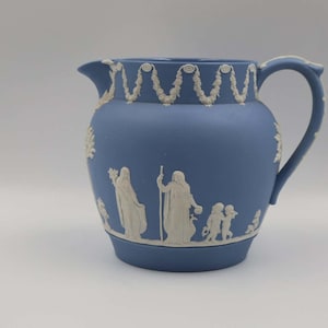 May include: A blue ceramic pitcher with a white relief design. The pitcher has a curved handle and a spout. The white relief depicts classical figures and decorative patterns. The pitcher is likely for decorative use.