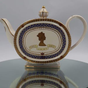 Könnte beinhalten: Cremefarbene Teekanne mit goldenen Verzierungen und einem dekorativen ovalen Feld mit einer Silhouette einer gekrönten Figur, den Jahren 1952 und 2002 sowie einer Krone. Das Feld hat einen blau-goldenen Musterrahmen. Die Teekanne hat einen gebogenen Griff und Ausguss.