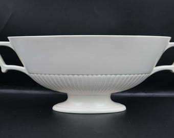 Może przedstawiać: Biała ceramiczna misa do serwowania z dwoma kwadratowymi uchwytami. Misa ma szeroki, płytki kształt z żłobioną dolną częścią i podstawą na cokole. Misa jest ustawiona na ciemnym tle.
