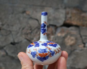 Puede incluir: Un pequeño florero de cerámica blanca con base redondeada y cuello delgado. La superficie está decorada con un patrón floral pintado a mano que presenta pétalos azules, acentos naranjas y finas líneas marrones en todo el exterior.