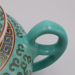 Puede incluir: Primer plano del asa y la tapa de una tetera de cer&aacute;mica turquesa. La tapa presenta un patr&oacute;n detallado con detalles en rojo, amarillo y azul. El asa tiene una forma suave y redondeada.