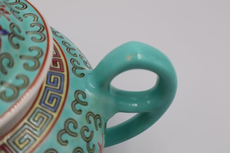 Puede incluir: Primer plano del asa y la tapa de una tetera de cer&aacute;mica turquesa. La tapa presenta un patr&oacute;n detallado con detalles en rojo, amarillo y azul. El asa tiene una forma suave y redondeada.