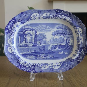 May include: An oval ceramic serving platter featuring a blue and white transferware pattern. The centre depicts a pastoral landscape with figures, trees, and architectural ruins, while the wide border displays intricate floral and geometric motifs.