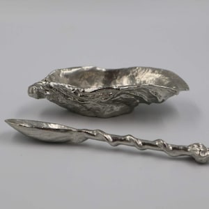 Puede incluir: Un pequeño cuenco plateado con un diseño orgánico texturizado, junto con una cuchara a juego. La cuchara tiene un mango retorcido y un extremo detallado. El conjunto se muestra sobre una superficie blanca.