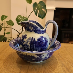 May include: A ceramic wash basin and matching pitcher set featuring a deep blue and white transferware pattern depicting detailed landscape scenes with buildings and trees. The pitcher has a curved handle and a wide spout, resting inside the bowl.