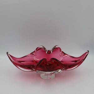 May include: A deep magenta glass bowl with a wide, elongated, and undulating rim that tapers to pointed ends. The bowl features a clear glass base and a smooth, translucent finish, showcasing a vibrant red-pink hue throughout the structure.