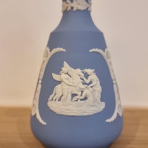 以下が含まれることがあります： 白のレリーフが施された青い花瓶。花瓶には、人物と翼のある馬が登場する古典的なシーンが描かれています。花瓶は、フレアベースと、装飾的な白いバンドが付いた細い首を持っています。