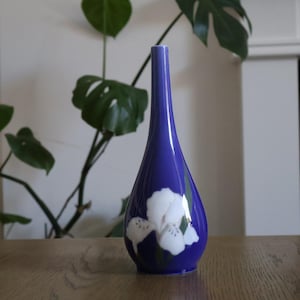 May include: A tall, teardrop-shaped ceramic vase with a smooth, deep blue glaze. The base of the vase features a painted white iris flower with subtle green leaves, extending upward from the bottom edge toward the center of the vessel body.
