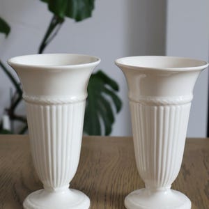 May include: Two off-white ceramic vases with a flared rim, a vertical ribbed body, and a circular pedestal base. Each vase features a decorative rope-like texture detail around the neck and the base, providing a consistent look for both pieces.