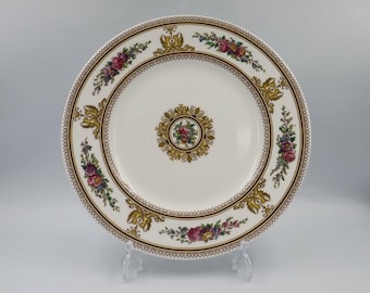 May include: A white ceramic plate with a decorative floral and gold rim. The plate features a central floral medallion and repeating floral and gold accents around the border. The plate is displayed on a clear stand.