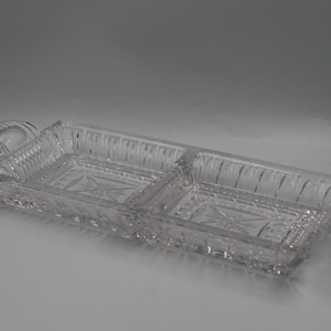 May include: A clear, rectangular glass serving dish featuring two divided compartments, two side handles, and intricate pressed patterns including starburst motifs at the base and vertical ribbed detailing along the outer walls.