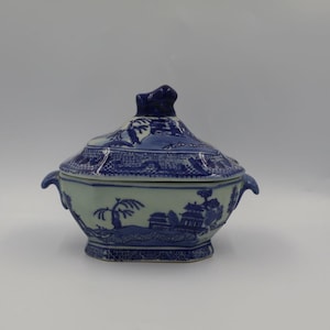 May include: A blue and white ceramic lidded serving dish. The dish features a detailed landscape design with trees and buildings. The lid has a decorative handle. A classic piece of tableware.