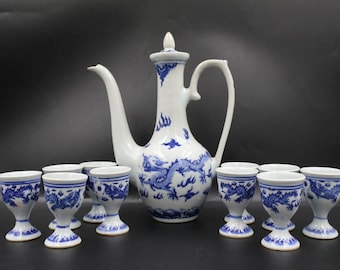 Puede incluir: Un juego de té de porcelana blanca con motivos de dragones azules. El juego incluye una tetera alta y ornamentada con asa y boquilla curvadas, y varias tazas pequeñas en forma de huevo. El diseño es de estilo clásico y tradicional.