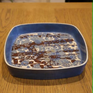 May include: A square ceramic dish with rounded corners and a solid blue exterior. The interior features a white base decorated with a repeating pattern of blue floral motifs and dark brown horizontal stripes that overlay the floral design.