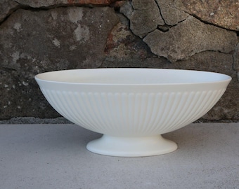 May include: A white, footed bowl with a fluted exterior design. The bowl has a wide, shallow shape and sits on a round base. The background features a textured stone wall.
