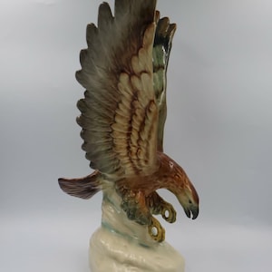 May include: A ceramic eagle figurine in a dynamic diving pose with wings fully extended upward. The bird features a glossy finish with shades of brown, tan, and cream, perched upon a textured, light-colored base that mimics a rocky formation.
