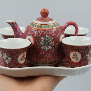 May include: A red and white ceramic tea set with a teapot, four cups, and a serving tray. The teapot and cups feature a floral design and Chinese characters. The set is held on a white tray.