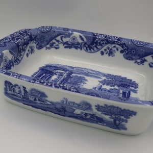 May include: A rectangular white ceramic baking dish featuring a blue transferware pattern. The design includes detailed landscape scenes with trees and architectural ruins on the exterior and interior, complemented by a floral border on the rim.