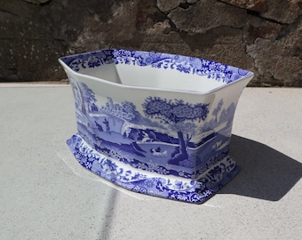 Puede incluir: Una jardinera rectangular de cerámica con base blanca y un detallado patrón azul de transferencia que representa un paisaje pastoral con árboles, figuras y animales. El borde y la base están decorados con un borde floral en azul a juego.