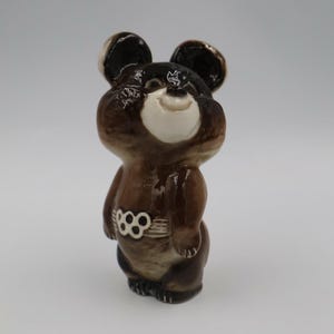May include: A glossy ceramic figurine of a brown bear with a white muzzle and ears. The bear has a smiling expression and wears a belt featuring a white five-ring Olympic symbol design around its midsection, rendered in a smooth, glazed finish.