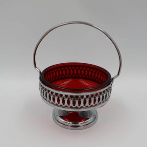 May include: A vintage red glass bowl with a decorative silver metal filigree base and handle. The bowl has a round shape and a detailed pattern around the rim. The handle arches over the top.