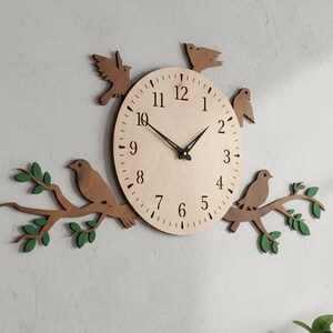 May include: A wooden wall clock with a light-colored face, black hands, and black numbers. The clock is surrounded by wooden birds and branches with green leaves. The clock is mounted on a white wall above a wooden cabinet.
