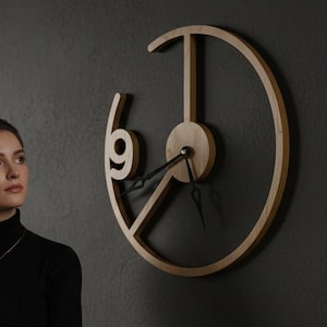 May include: A modern, minimalist wall clock with a light wood finish. The clock features a unique open design with the number 9 prominently displayed. The hands are black, and the clock is mounted on a dark gray wall. The clock's diameter is approximately 18 inches.