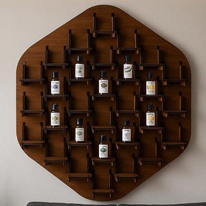 Puede incluir: Un estante de pared hexagonal de madera marrón oscuro con múltiples estantes pequeños, que exhibe varias botellas de aceites esenciales. La unidad está montada en una pared de color claro sobre un sofá gris. Una planta alta en maceta está a la izquierda.