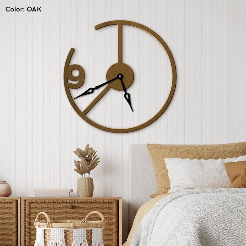 May include: Oak-colored wall clock with a unique open design. The clock features a circular frame with the number 9 and black clock hands. The text "Color: OAK" is in the upper left corner. The clock is mounted on a white wall.