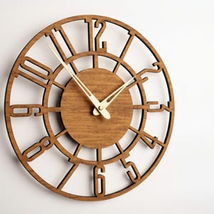 Puede incluir: Un reloj de madera redondo con una esfera marr&oacute;n claro y un marco marr&oacute;n oscuro. El reloj presenta n&uacute;meros romanos recortados y manecillas de color claro. El reloj est&aacute; montado en una pared blanca.