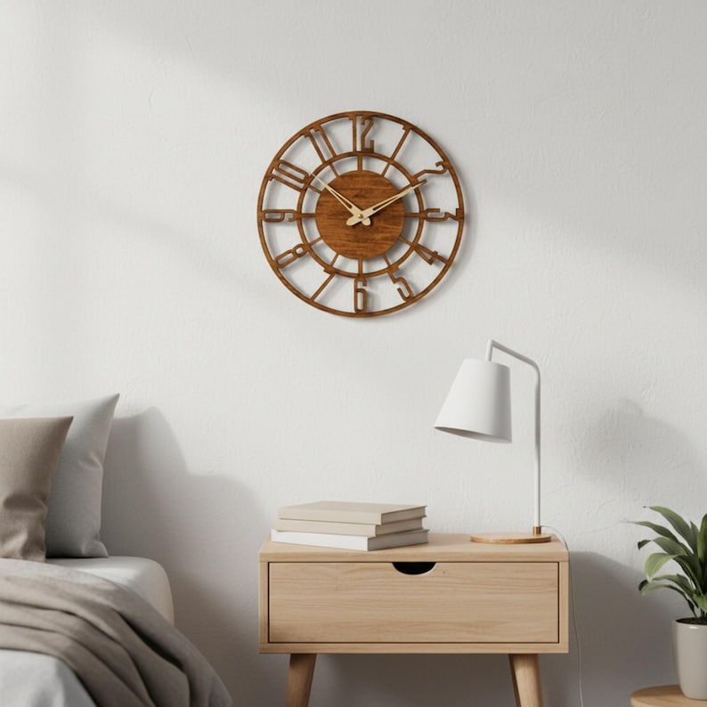 Puede incluir: Un reloj de pared redondo de madera con acabado marr&oacute;n oscuro. El reloj presenta n&uacute;meros romanos y un dise&ntilde;o circular. Est&aacute; colgado en una pared blanca. Las manecillas del reloj son doradas. El reloj est&aacute; encima de una mesita de noche de madera con una l&aacute;mpara y libros.