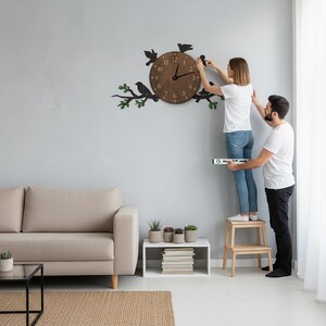 May include: A brown wooden wall clock with black bird and branch accents. The clock face has black numerals and hands. The clock is being installed on a light gray wall. The clock is approximately 12 inches in diameter.