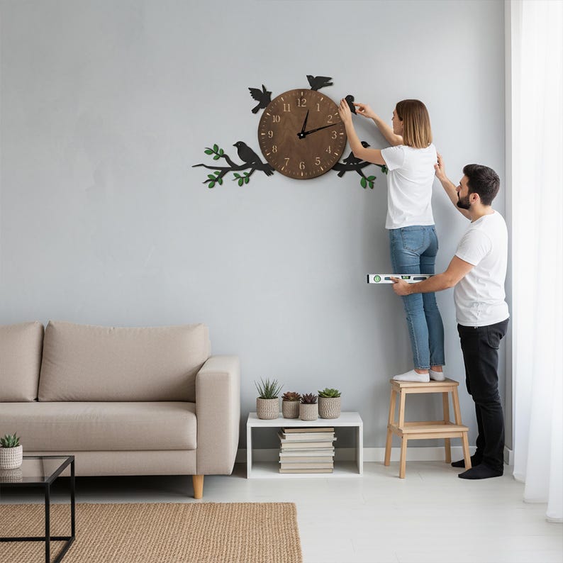 May include: A brown wooden wall clock with black bird and branch accents. The clock face has black numerals and hands. The clock is being installed on a light gray wall. The clock is approximately 12 inches in diameter.
