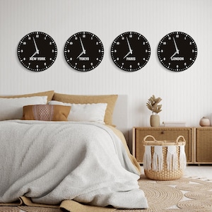 May include: Four black wall clocks showing the time in New York, Tokyo, Paris, and London. Each clock has white numerals and hands. The clocks are mounted on a white wall above a bed, with a neutral colour scheme.