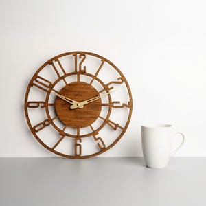 Puede incluir: Un reloj de madera redondo con acabado marr&oacute;n oscuro. La esfera del reloj presenta n&uacute;meros recortados y un c&iacute;rculo central. Las manecillas doradas indican la hora. Una taza blanca est&aacute; a la derecha del reloj.