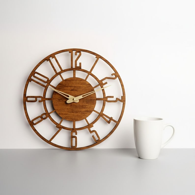Puede incluir: Un reloj de madera redondo con acabado marr&oacute;n oscuro. La esfera del reloj presenta n&uacute;meros recortados y un c&iacute;rculo central. Las manecillas doradas indican la hora. Una taza blanca est&aacute; a la derecha del reloj.