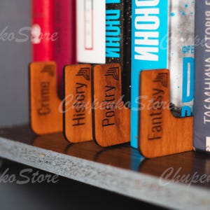 May include: Five wooden bookends with different categories engraved on them: Crime, History, Poetry, Fantasy, and a bookend with a blank space. The bookends are brown and have a natural wood finish. The bookends are on a shelf with books behind them.