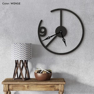 May include: A black metal wall clock with a minimalist design, featuring a partial circular frame and the number 9. The clock hands are black. A wooden table holds a lamp with a gray and white chevron pattern shade and a decorative bowl.