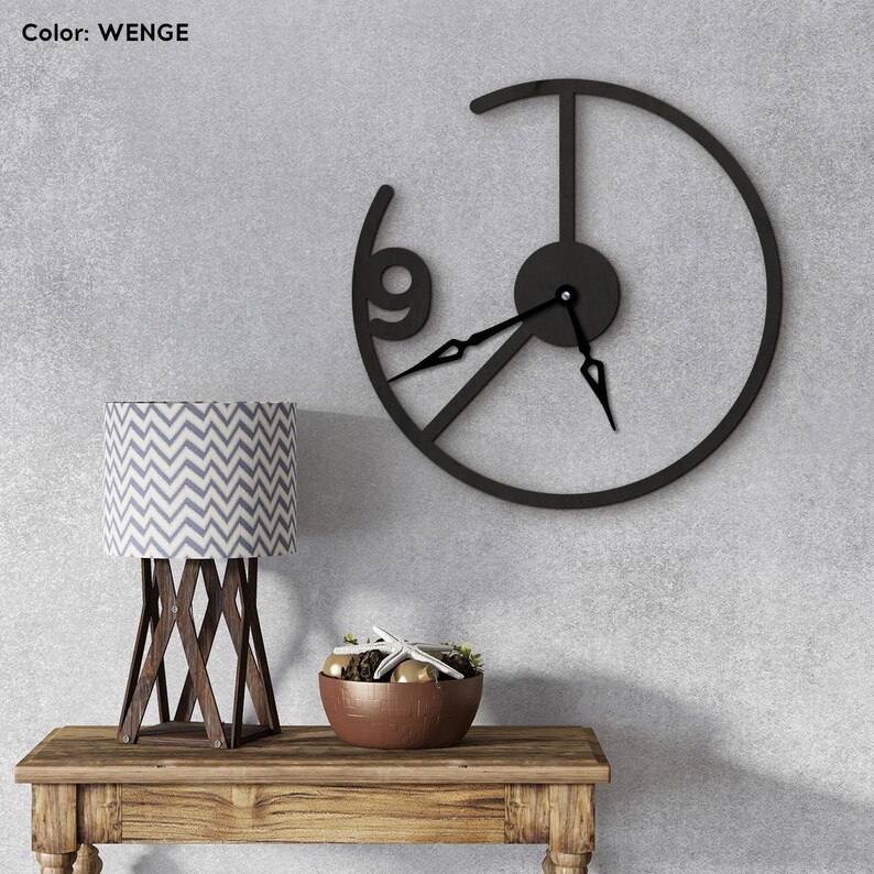 May include: A black metal wall clock with a minimalist design, featuring a partial circular frame and the number 9. The clock hands are black. A wooden table holds a lamp with a gray and white chevron pattern shade and a decorative bowl.