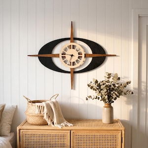 Pode incluir: Um relógio de parede com um mostrador circular claro com raios, montado sobre uma ponta de madeira castanha horizontal e uma grande moldura oval preta. O relógio tem dois ponteiros pretos finos e está posicionado numa parede branca com painéis.