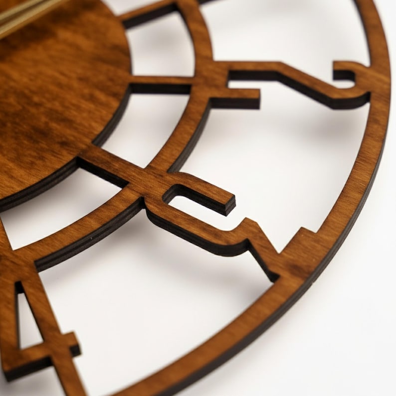 Puede incluir: Primer plano de una esfera de reloj de madera con un acabado marr&oacute;n oscuro. El reloj presenta un dise&ntilde;o recortado con n&uacute;meros romanos y una forma circular. Las manecillas son visibles, indicando la hora. El reloj es un art&iacute;culo decorativo.