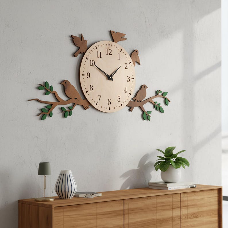 May include: A wooden wall clock with a light-colored face, black hands, and black numbers. The clock is surrounded by wooden birds and branches with green leaves. The clock is mounted on a white wall above a wooden cabinet.