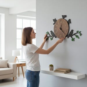 May include: A wooden wall clock with a brown face and black hands, surrounded by black bird silhouettes and green leaves. The clock is being mounted on a gray wall. The clock's face has the numbers 1 through 12.