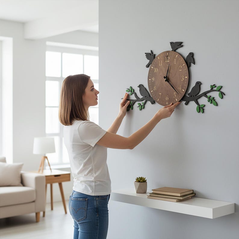 May include: A wooden wall clock with a brown face and black hands, surrounded by black bird silhouettes and green leaves. The clock is being mounted on a gray wall. The clock's face has the numbers 1 through 12.