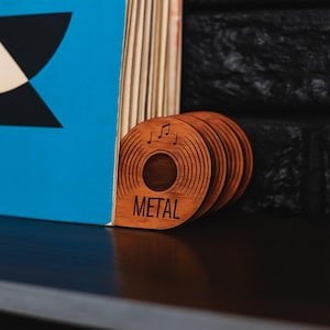 Może przedstawiać: Drewniane podstawki w kształcie płyt winylowych. Brązowe podstawki mają wytrawione rowki i nuty. Słowo "METAL" jest wytrawione na przodzie. Są ułożone obok niebieskiej książki.