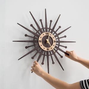 May include: A mid-century modern wall clock with a starburst design. The clock features a dark brown wooden frame with radiating spikes and a light-colored circular face. The clock hands are gold-colored.