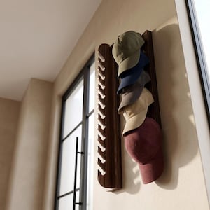 Puede incluir: Un perchero de madera para sombreros montado en la pared, con seis gorras de béisbol de varios colores. Las gorras incluyen tonos de verde, azul, gris, beige y burdeos. El perchero tiene un acabado de madera oscura.