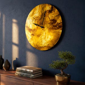 May include: A round wall clock with a textured, golden-yellow face and black hands. The clock is mounted on a dark blue wall. The clock's diameter is approximately 12 inches. Decorative items, including books and a bonsai tree, are on a wooden surface below.